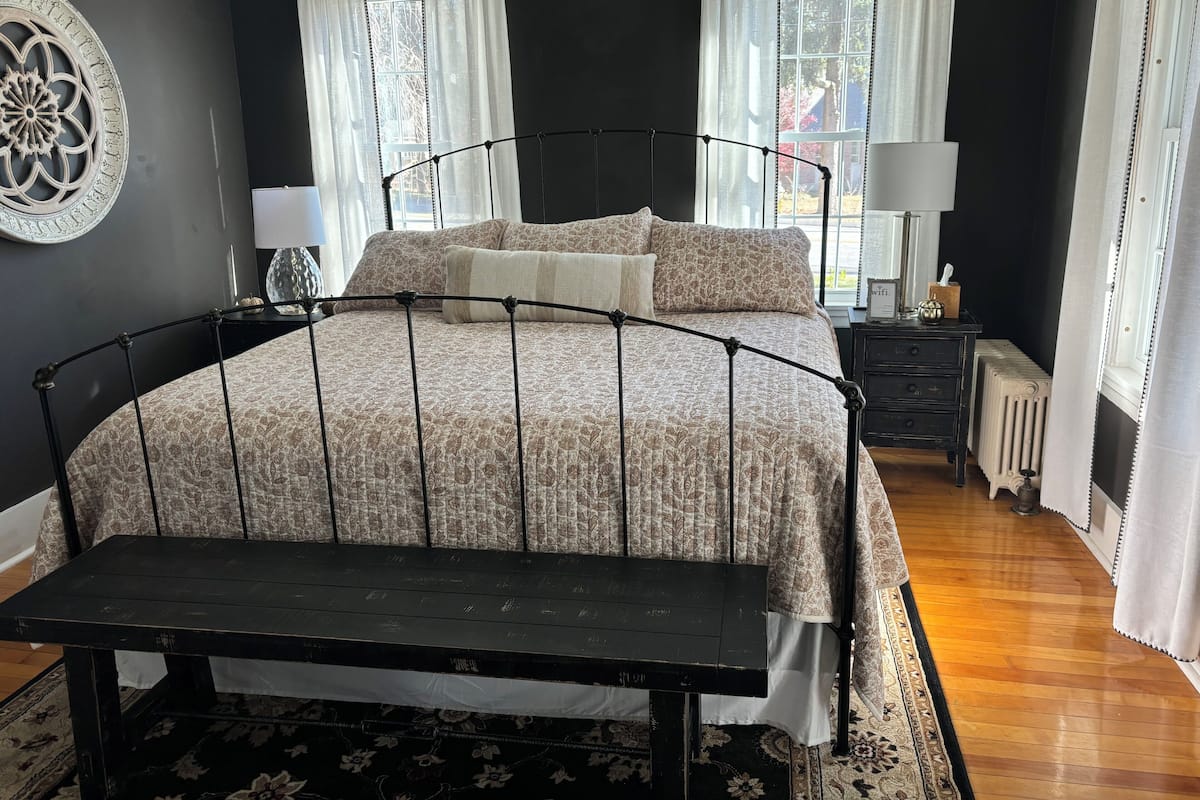 A bedroom with a bed, a bench, a nightstand, a lamp, and a radiator.