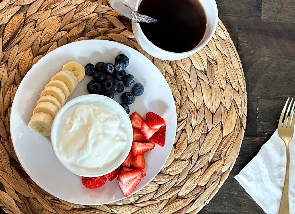 Une assiette contenant du yaourt, des tranches de bananes, des myrtilles et des fraises, une tasse de café et un set de table en fibres tressées.