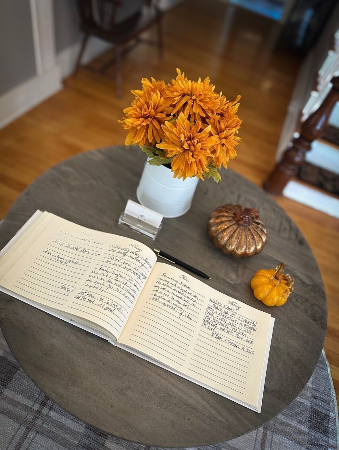 Une table ronde avec un carnet, un stylo, un petit vase blanc contenant des fleurs orange et deux petites courges.