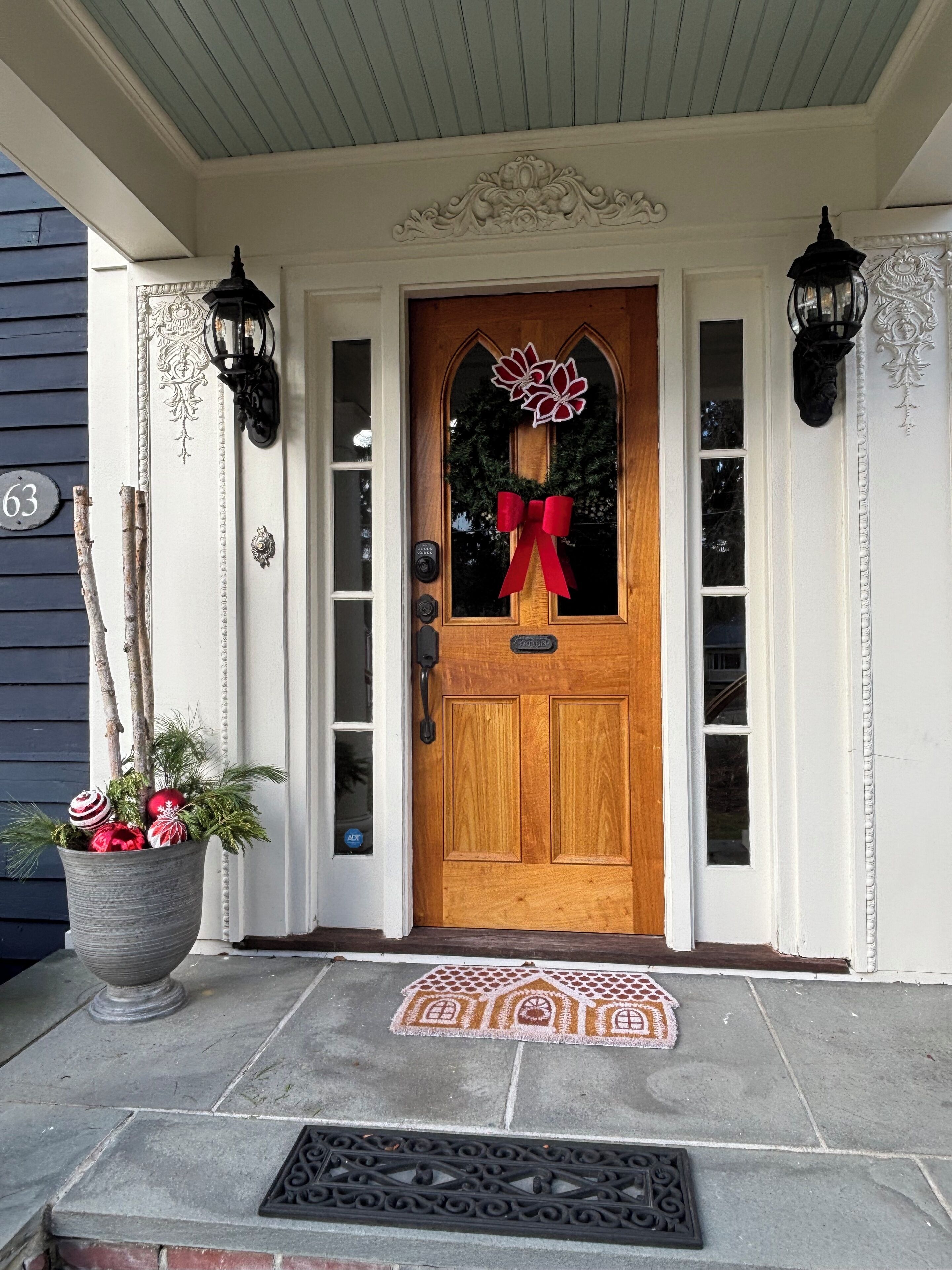A wooden front door with a red bow, a potted plant with greenery and ornaments, and a doormat with a house design.