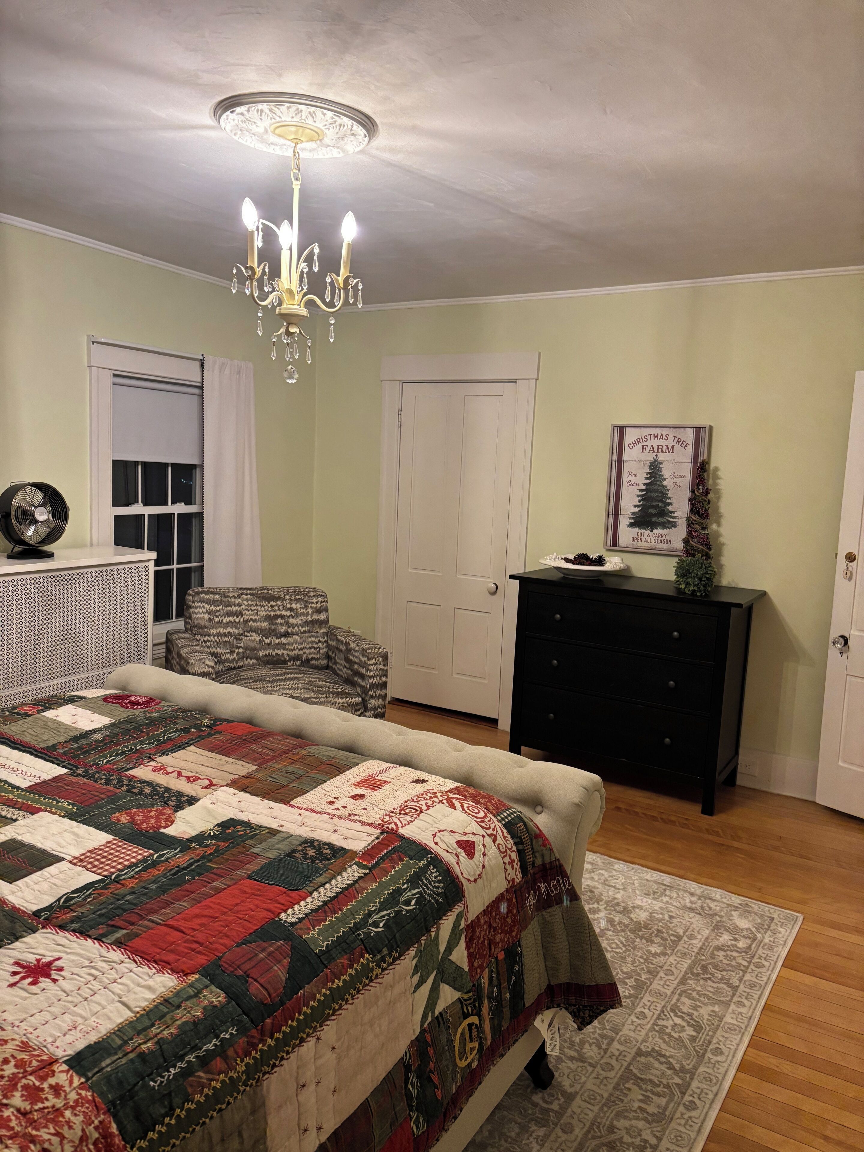 A bedroom with a large bed, a chair, a dresser, a fan, a chandelier, and a door.