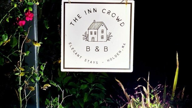 Un letrero para The Inn Crowd B&B con una ilustración de una casa, que dice "Estancias Elegantes - Holden, MA".