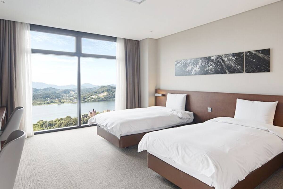 Habitación de hotel con dos camas, un escritorio y un amplio ventanal con vistas a un lago y montañas.
