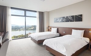 A hotel room with two beds, a desk, and a large window offering a view of a lake and mountains.