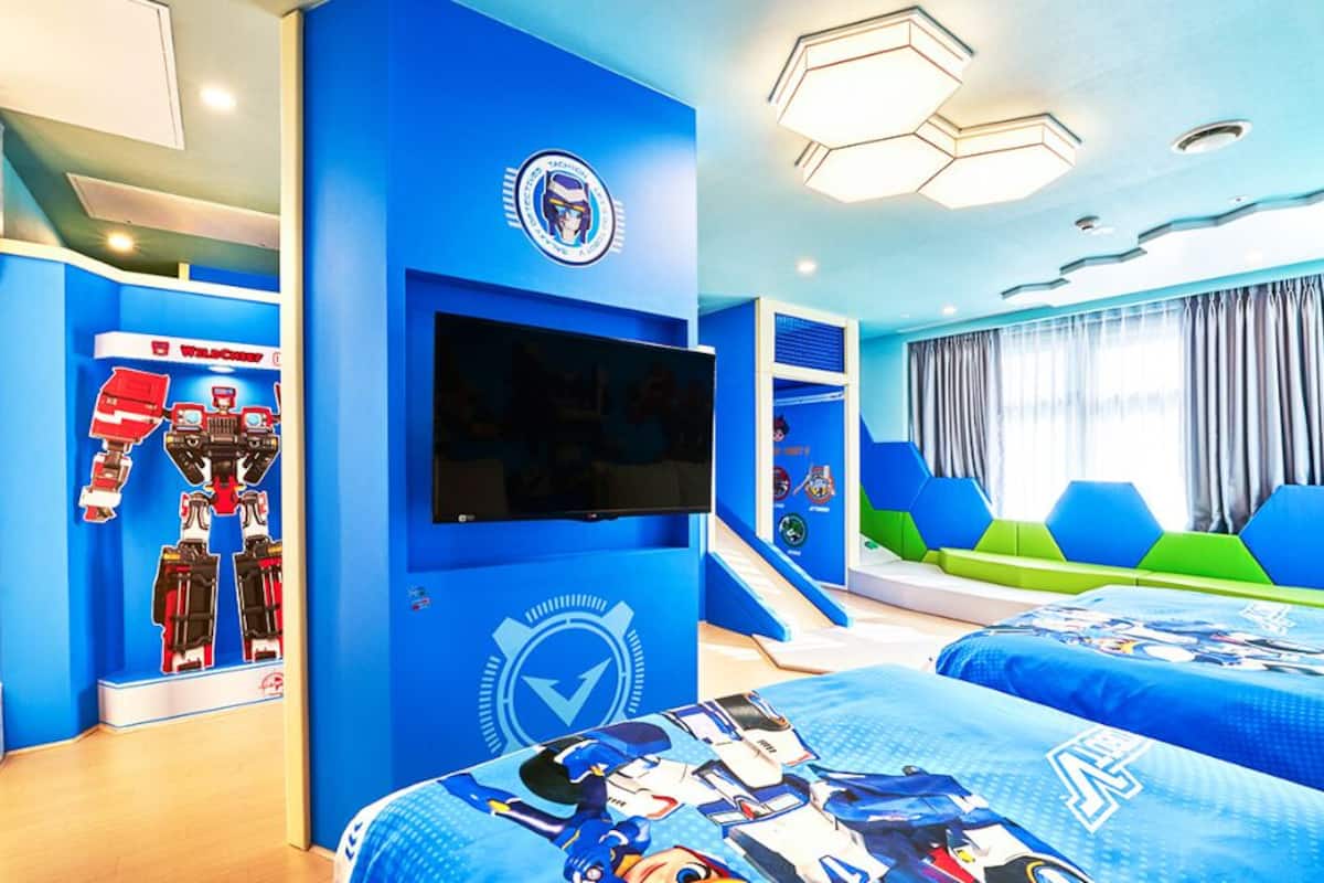 Una habitación con paredes azules, un televisor montado en la pared, una cama con colcha azul y blanca, y una ventana con cortinas.