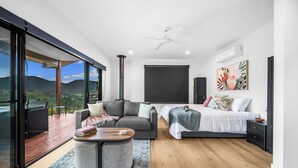 A modern bedroom with a large bed, a sofa, and a coffee table. There is a sliding glass door leading to an outdoor area with a view of the mountains.