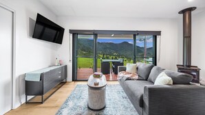 A modern living room with a sofa, a coffee table, and a TV mounted on the wall. There is a sliding glass door leading to an outdoor area with a table and chairs.