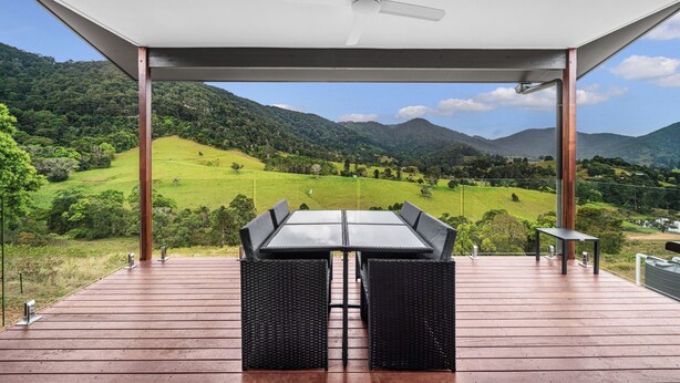 A covered outdoor seating area with a table and chairs, overlooking a green valley and mountains.
