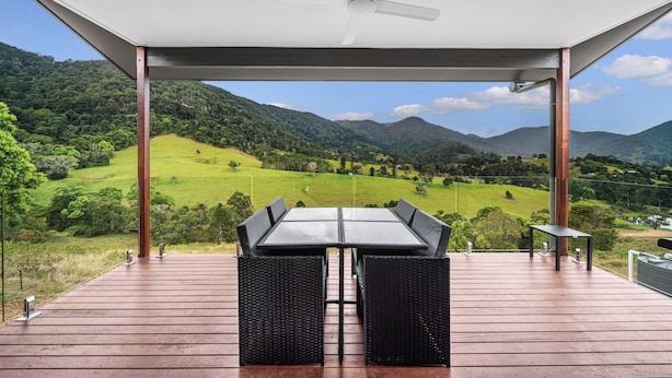 A covered outdoor seating area with a table and chairs, overlooking a green valley and mountains.