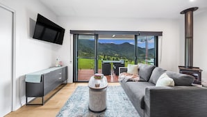 A modern living room with a sofa, a coffee table, and a TV mounted on the wall. There is a sliding glass door leading to an outdoor area with a table and chairs.
