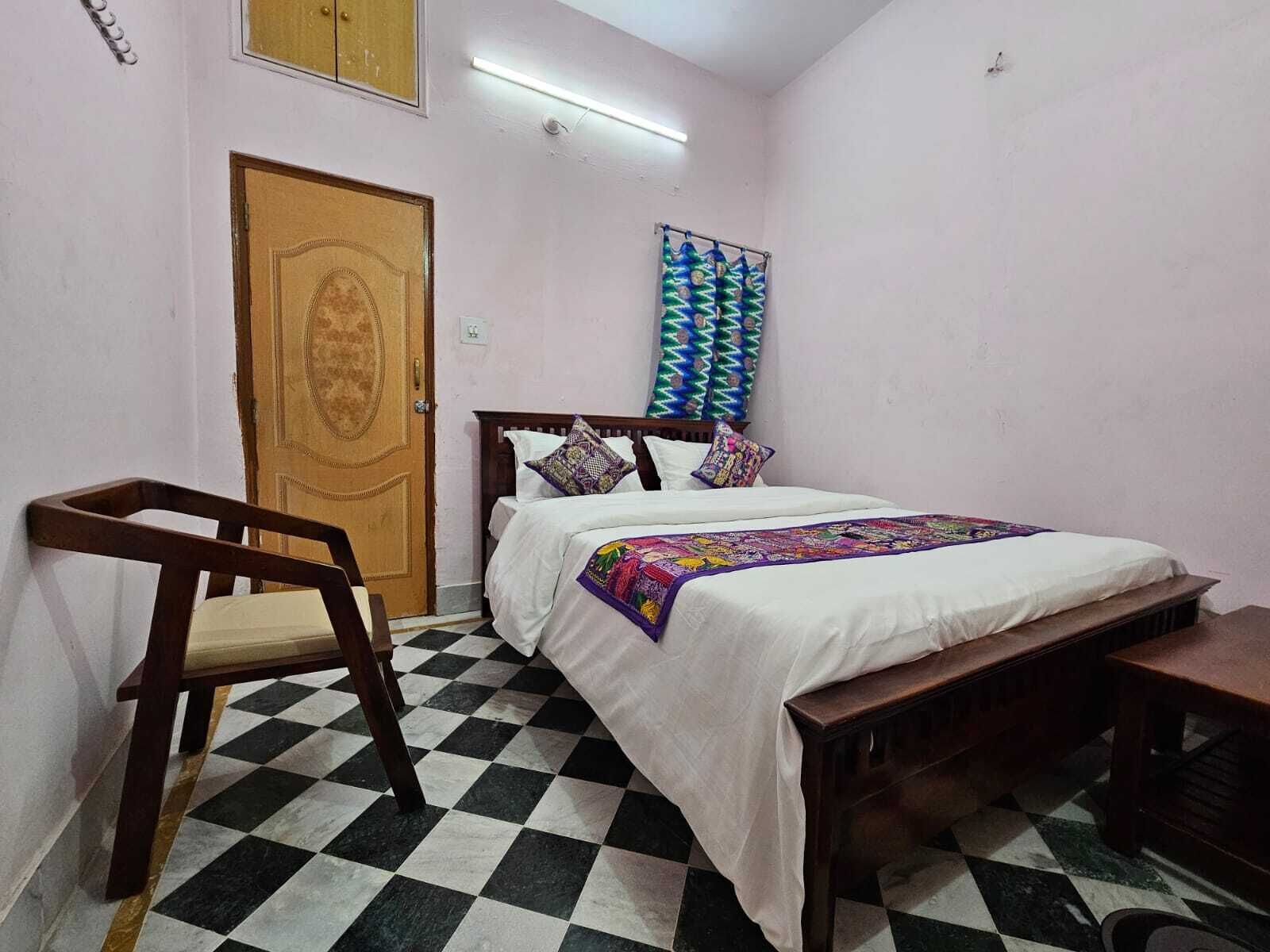 Un dormitorio con piso a cuadros, una cama con colcha colorida, una silla de madera, un banco y una puerta con panel decorativo.