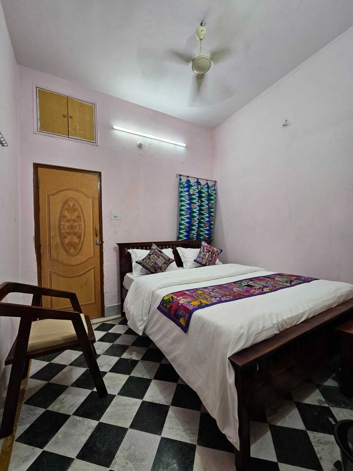 Un dormitorio con cama, una silla de madera, piso a cuadros, ventilador de techo y una puerta.