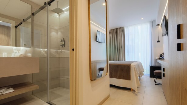 Ein modernes Hotelzimmer mit einer Duschkabine aus Glas, einem großen Spiegel, einem Bett mit brauner Decke, einem kleinen Hocker und einem an der Wand montierten Fernseher.