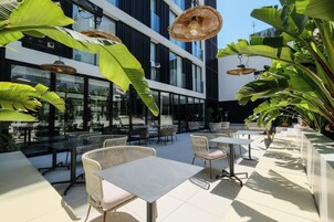 Un espacio al aire libre con mesas y sillas, rodeado de plantas verdes grandes y lámparas de mimbre colgantes.