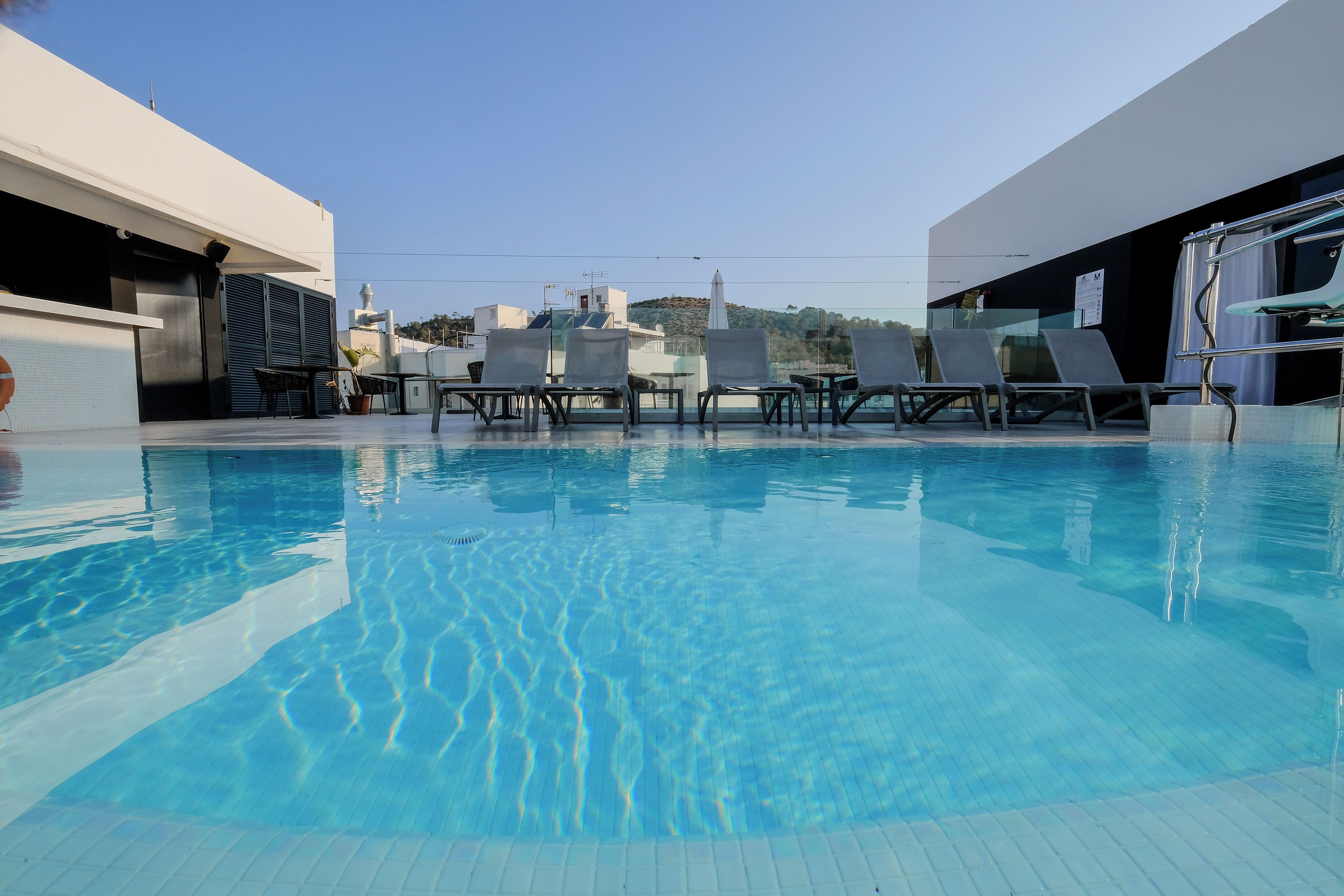 A rooftop swimming pool with lounge chairs and a table, surrounded by modern buildings and a clear sky.