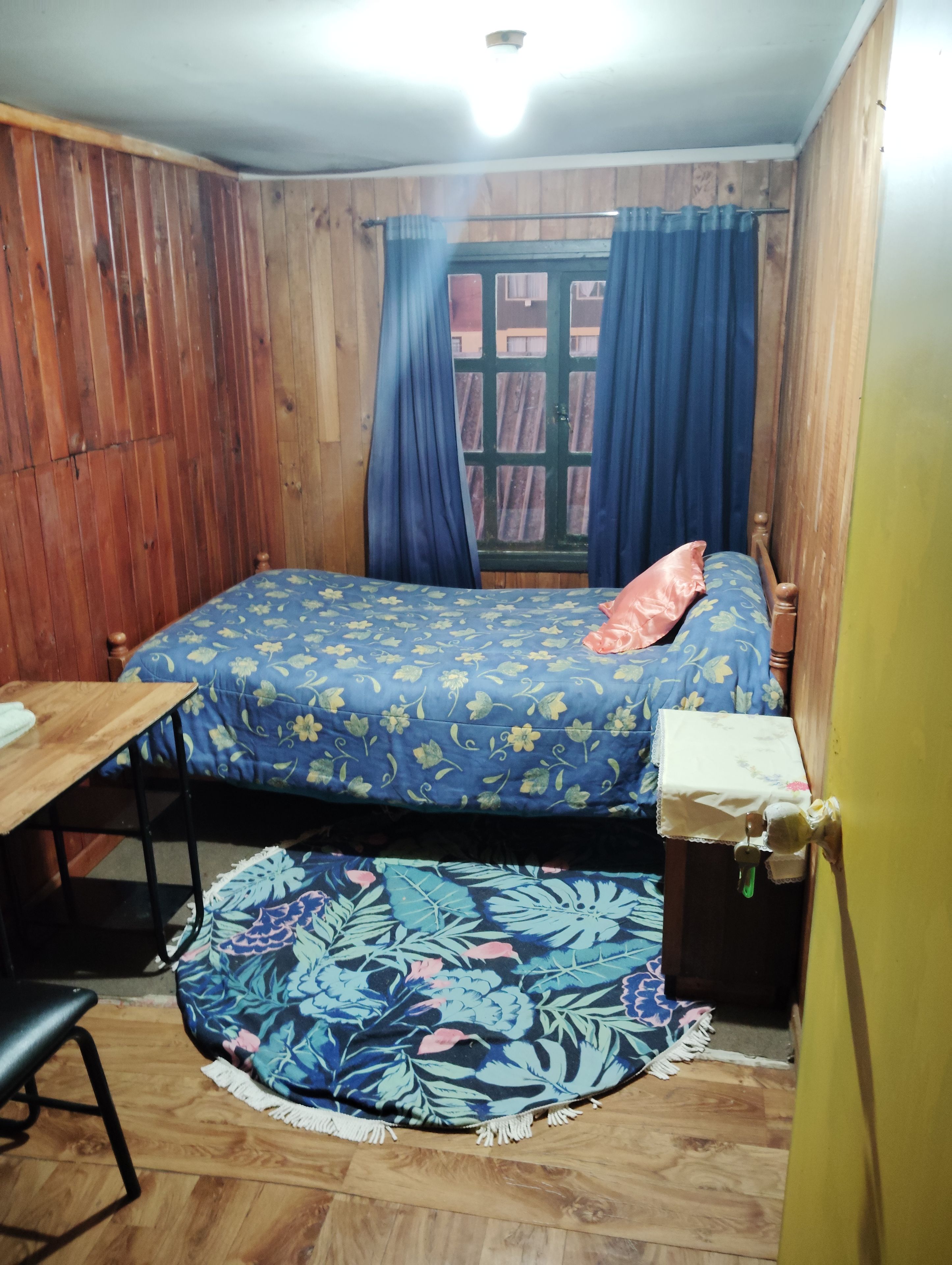 A small, wooden-furnished room with a bed, a table, and a chair.