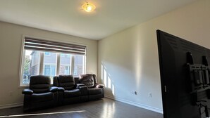 A spacious living room with a large sectional sofa, a flat-screen TV, and a large window with blinds. - Newly Built Detached Home! Close to nature and city.  (Brampton)