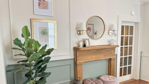 A hallway with a white wall, a wooden mantelpiece, two pink ottomans, a potted plant, and framed artwork.