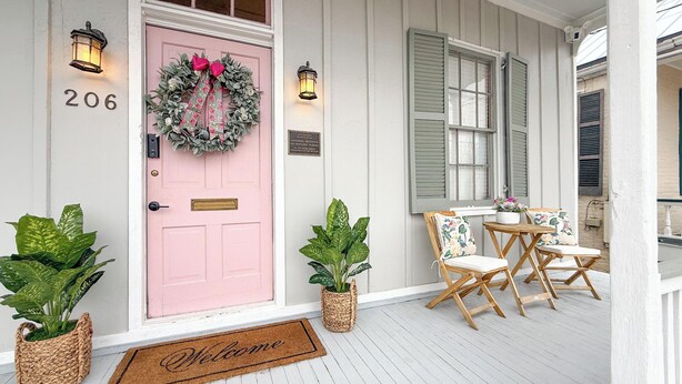 A porch with a pink door, two potted plants, a welcome mat, and a table with chairs.