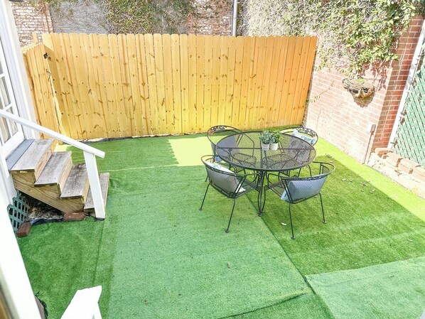 Un patio con césped artificial, una mesa redonda de metal y cuatro sillas.