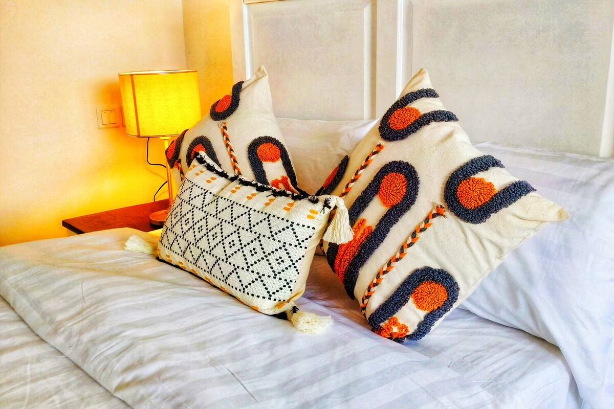 A bed with white linens, two decorative pillows with geometric patterns, and a woven bag on the side.