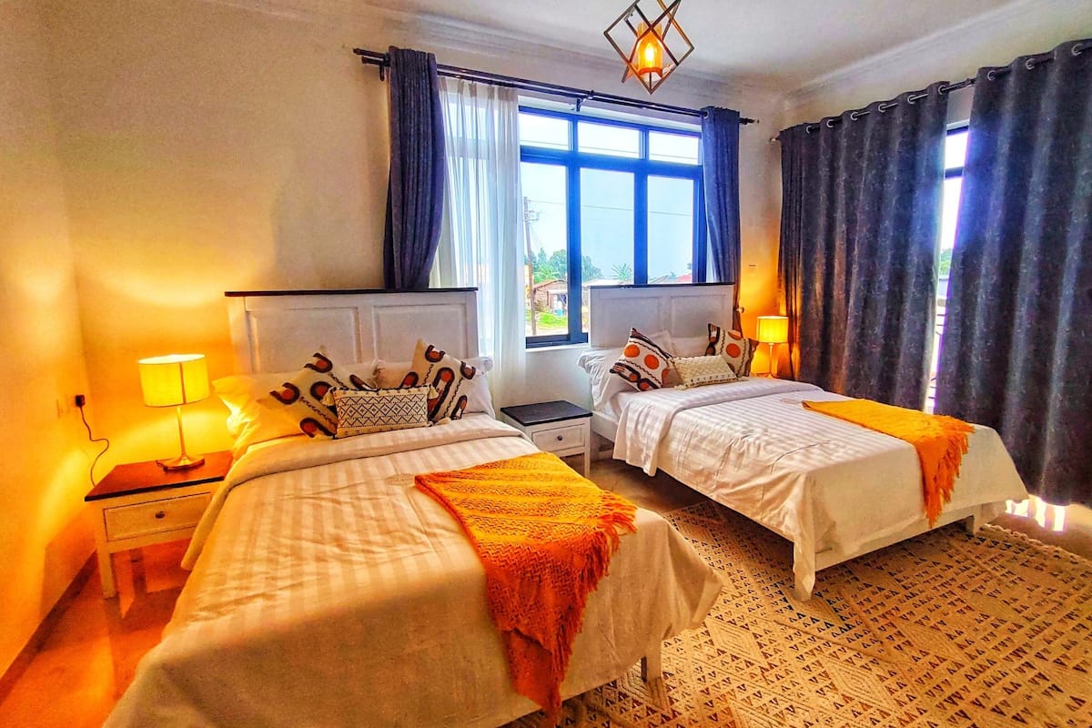 A hotel room with two beds, a window with curtains, a nightstand with a lamp, and a patterned rug.