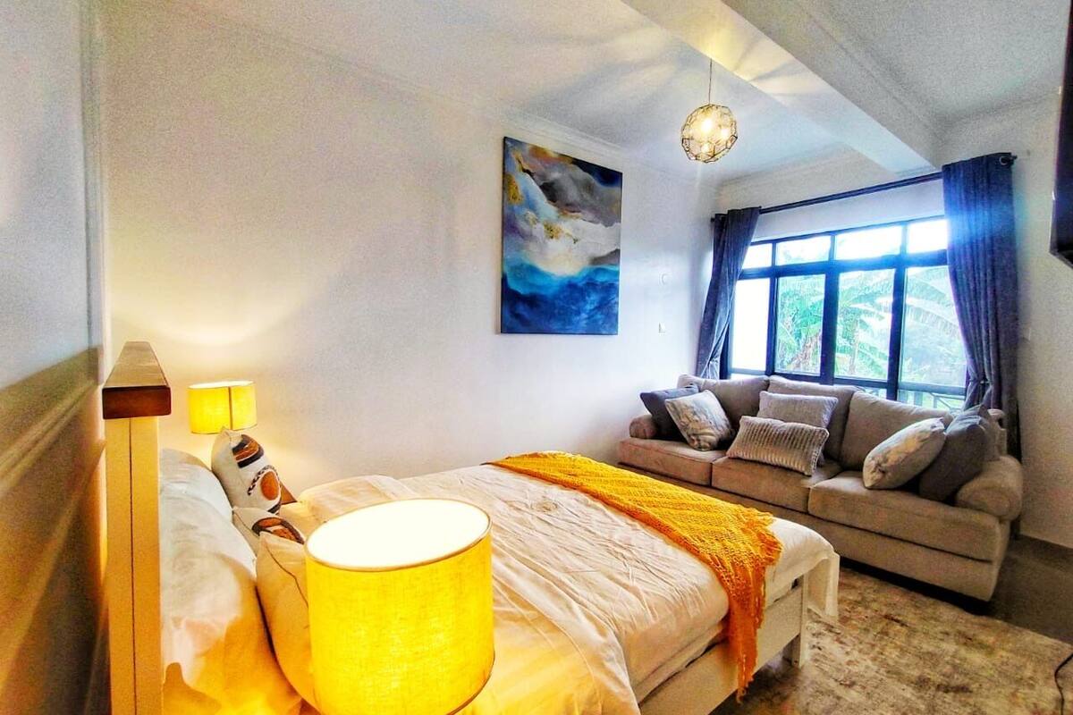 A bedroom with a bed, bedside lamps, a sofa, a window with curtains, and a painting on the wall.