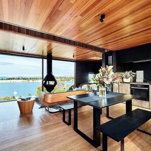 A modern kitchen with a large island, a dining area with a bench, and a view of a body of water. - The Tailored Pod - Holiday Houses Tasmania (Lewisham)