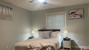 Ein Schlafzimmer mit einem Bett, zwei Nachttischen, einem Deckenventilator, einem Fenster mit Jalousien und Bildern an den Wänden.