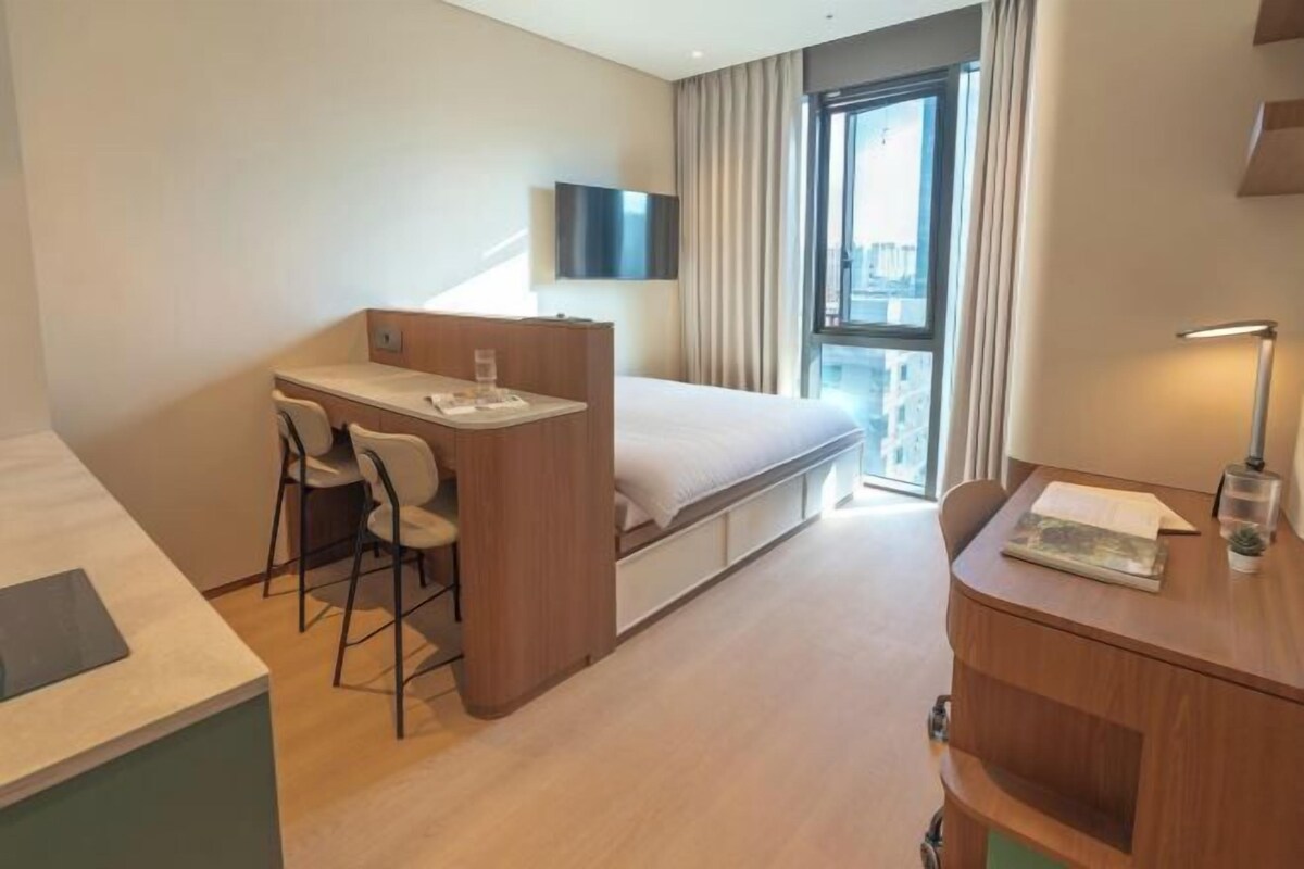 A compact hotel room with a bed, a desk with two chairs, a TV, and a window with curtains.