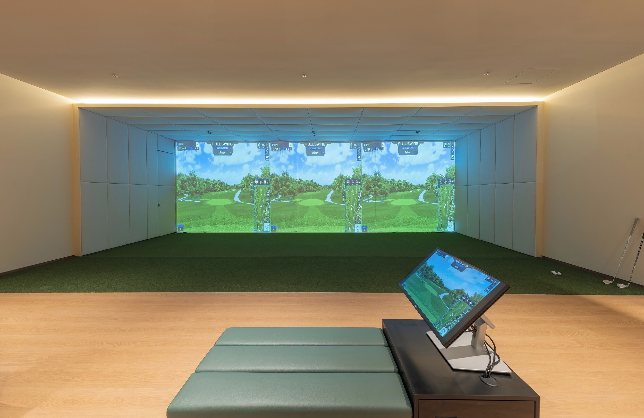 Una habitación con una pantalla grande mostrando una simulación de campo de golf, un monitor pequeño sobre un soporte y un suelo cubierto de alfombra verde.