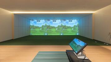 Una habitación con una pantalla grande mostrando una simulación de campo de golf, un monitor pequeño sobre un soporte y un suelo cubierto de alfombra verde.