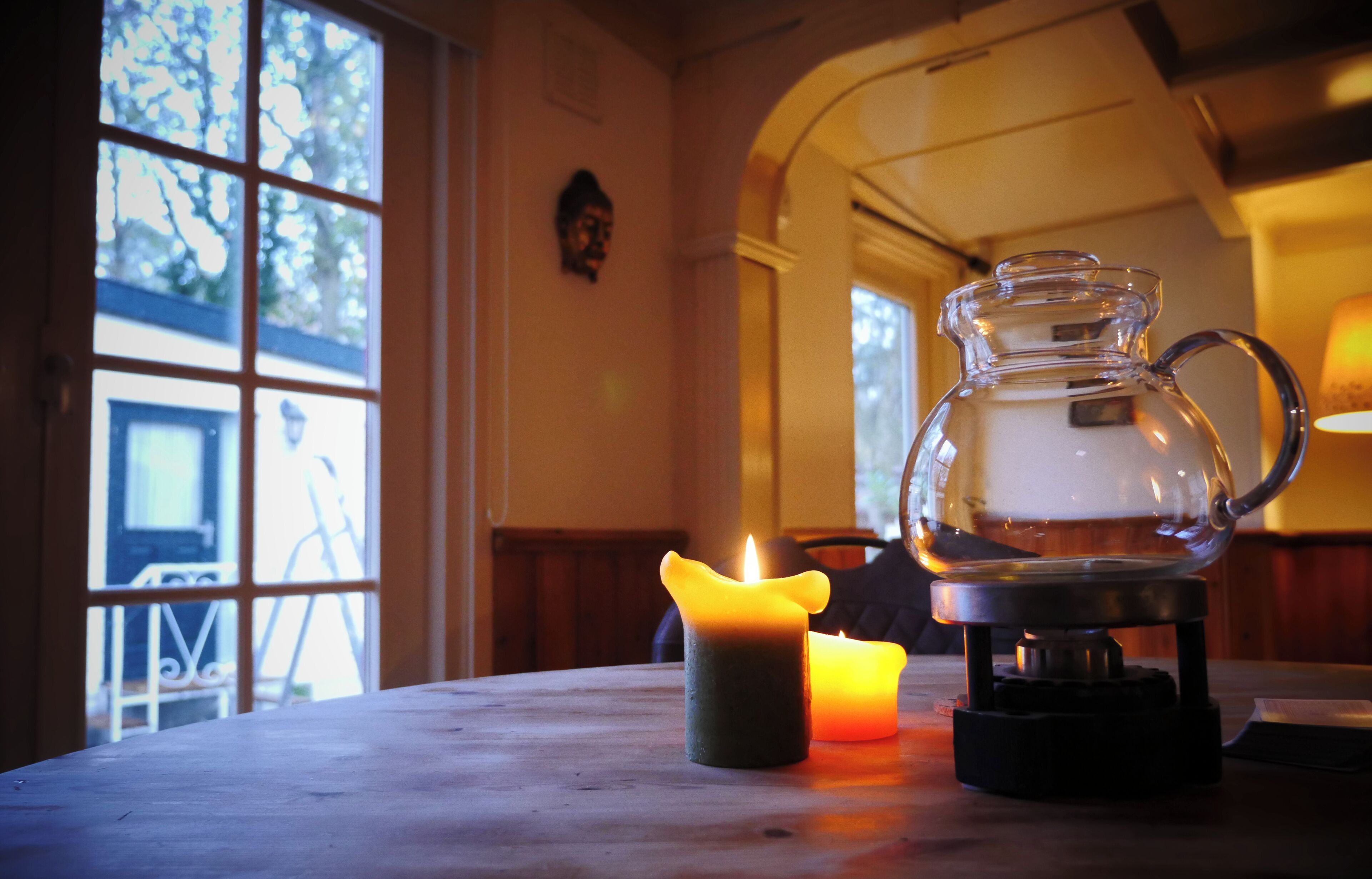 Un tavolo con una caraffa di vetro e due candele, una finestra vista alberi e una lampada sullo sfondo.