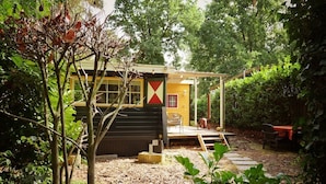 A small, single-story building with a yellow and black exterior, surrounded by dense greenery. - Cosy Cabin in the Woods - Perfect Get Away in a peaceful and green setting (Diessen)