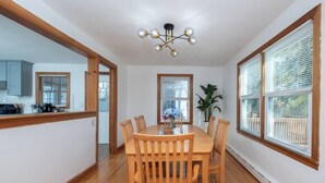 Un comedor con una mesa y sillas de madera, una araña, ventanas amplias y vistas al exterior.