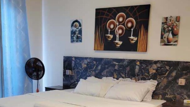 A neatly made bed with a white comforter and pillows, a headboard with a marble-like pattern, a wall-mounted artwork, and a small bedside table with a lamp. - Remarkable 3-Bed House in Kigali (Kigali)