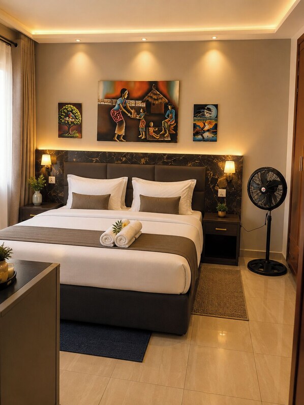 A hotel room with a large bed, two bedside tables, a standing fan, and artwork on the walls.