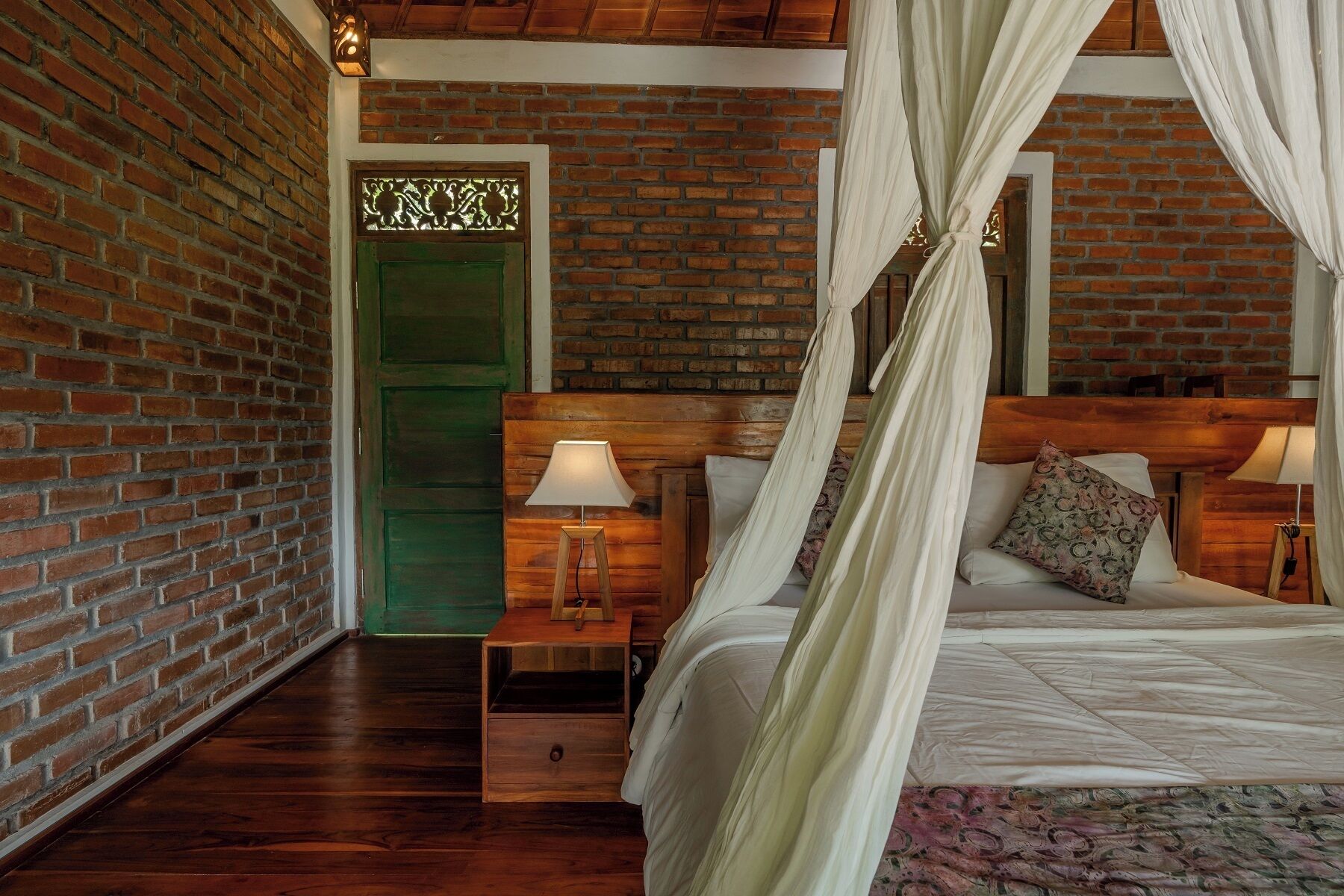 A bedroom with a brick wall, a green door, a wooden headboard, a bed with white bedding, a nightstand with a lamp, and a wooden bench.