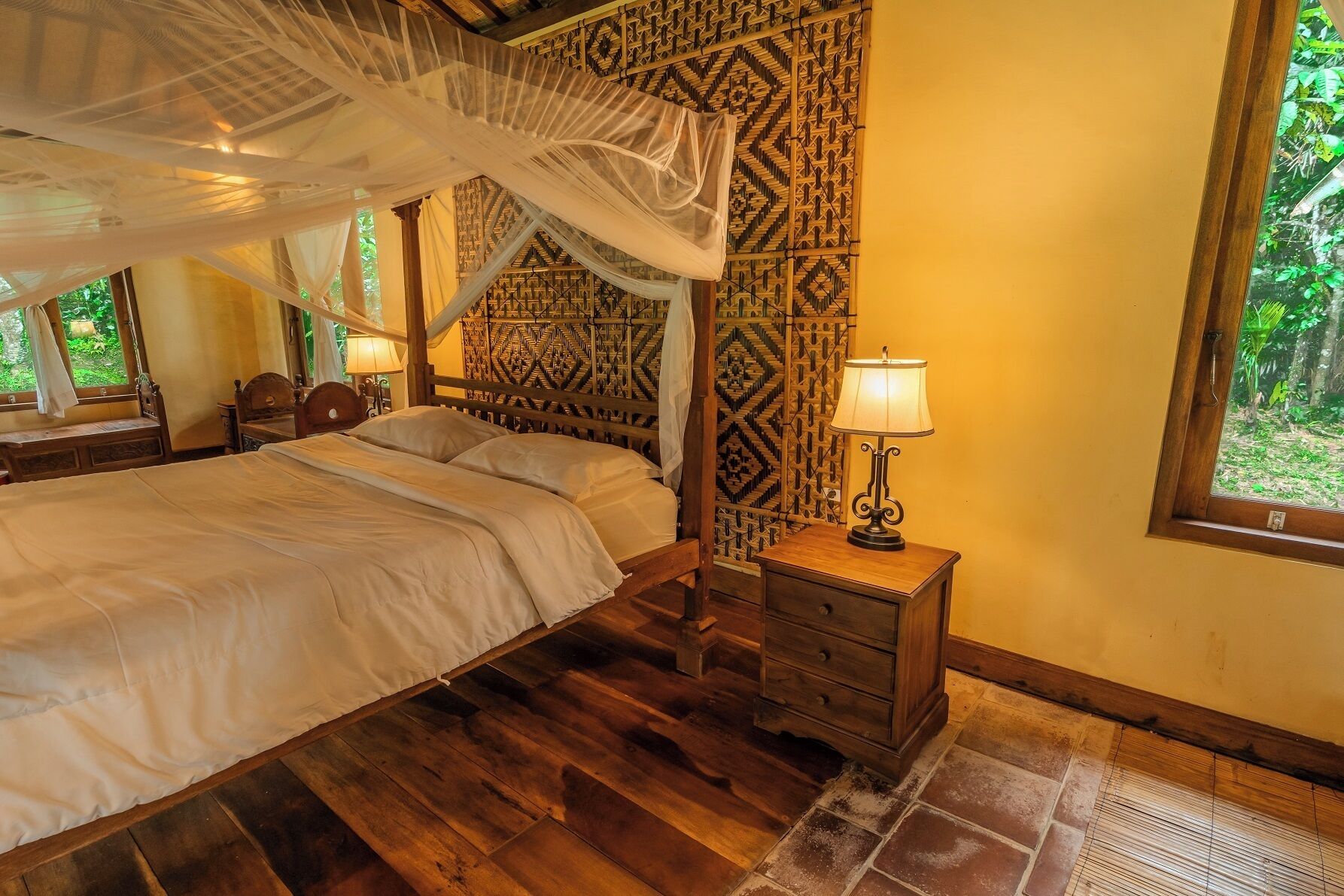 Un dormitorio con cama con dosel, una mesita de noche con lámpara, decoración de pared con estampado y una ventana con vistas a zonas verdes.