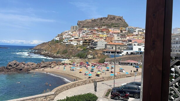 Un pueblo costero con una fortaleza histórica en una colina, una playa arenosa con sombrillas y una vista despejada del océano.