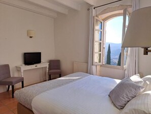 A hotel room with a bed, a desk with a television, a chair, and a window with a view of trees. - Logis Hotel Le Clos Des Sullys (Montclus)