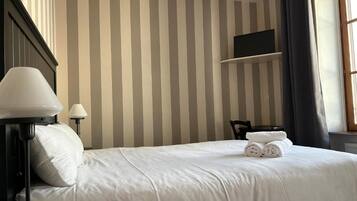 Habitación de hotel con pared a rayas, una cama con ropa de cama blanca, un escritorio con lámpara y un televisor en un estante.