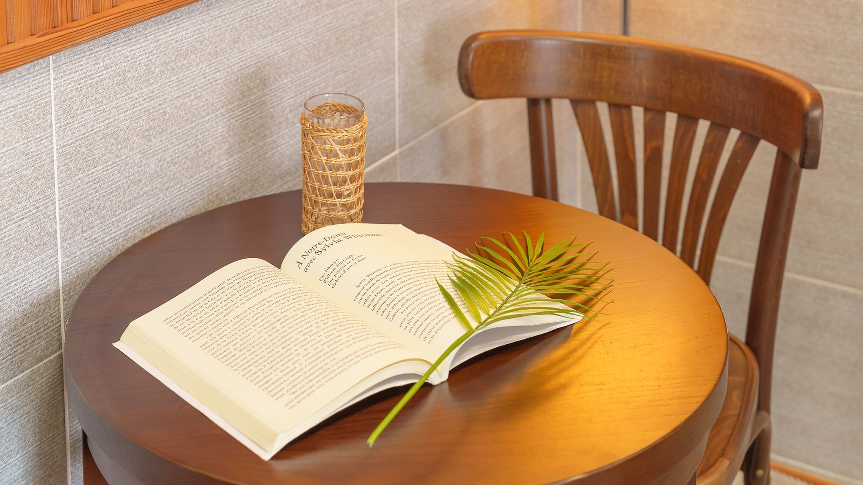 Un llibre obert sobre una taula de fusta, amb una fulla de palmera i un portaveus de vidre trenat.