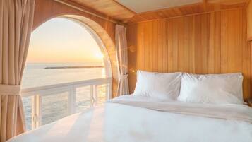 A bedroom with a large window overlooking a body of water and a distant structure, wooden paneling on the walls, and a bed with white bedding.