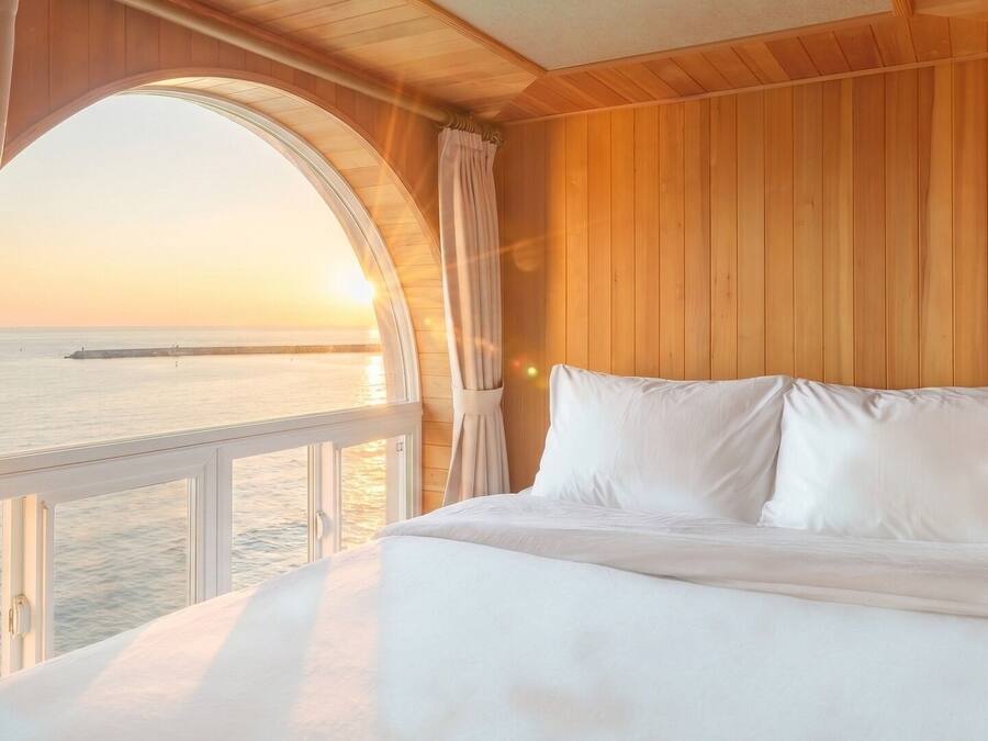 A bedroom with a large window overlooking a body of water and a distant structure, wooden paneling on the walls, and a bed with white bedding.