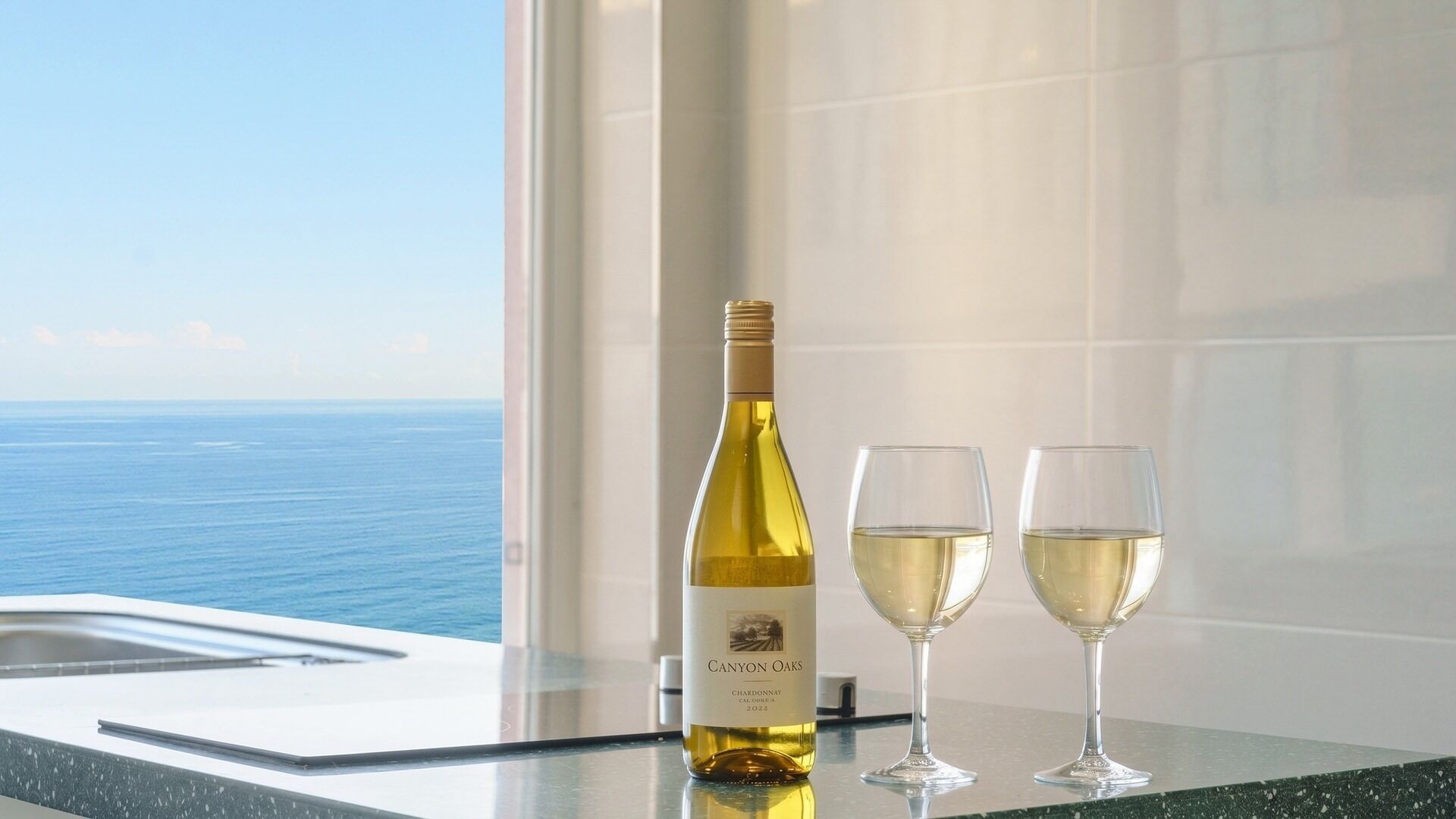 Una botella de Chardonnay Canyon Oaks 2022 y dos copas sobre una superficie reflectante, con vista al océano a través de una ventana.