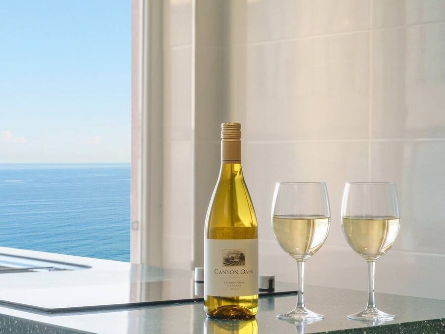 Una botella de Chardonnay Canyon Oaks 2022 y dos copas sobre una superficie reflectante, con vista al océano a través de una ventana.