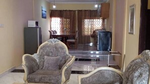 A living room with two ornate armchairs, a small table, a dining area with a table and chairs, and a kitchen area with a refrigerator and cabinets.