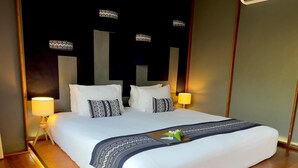 Ein ordentlich bezogenes Bett mit weißen Leinen und einer dunkel gemusterten Decke, zwei Nachttische mit Lampen und ein modernes Kopfstück mit vertikalen Leuchten.