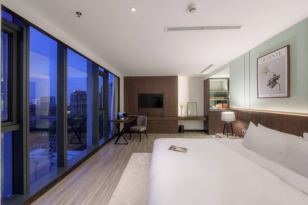 A modern hotel room with a large bed, a desk with a chair, a television, and a large window offering a city view.